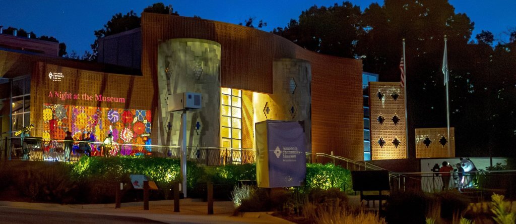 The Smithsonian Anacostia Community Museum's entrance is lit up with festive lighting that reads "A Night at the Museum" for an evening event. Behind it the sky is cobalt blue.