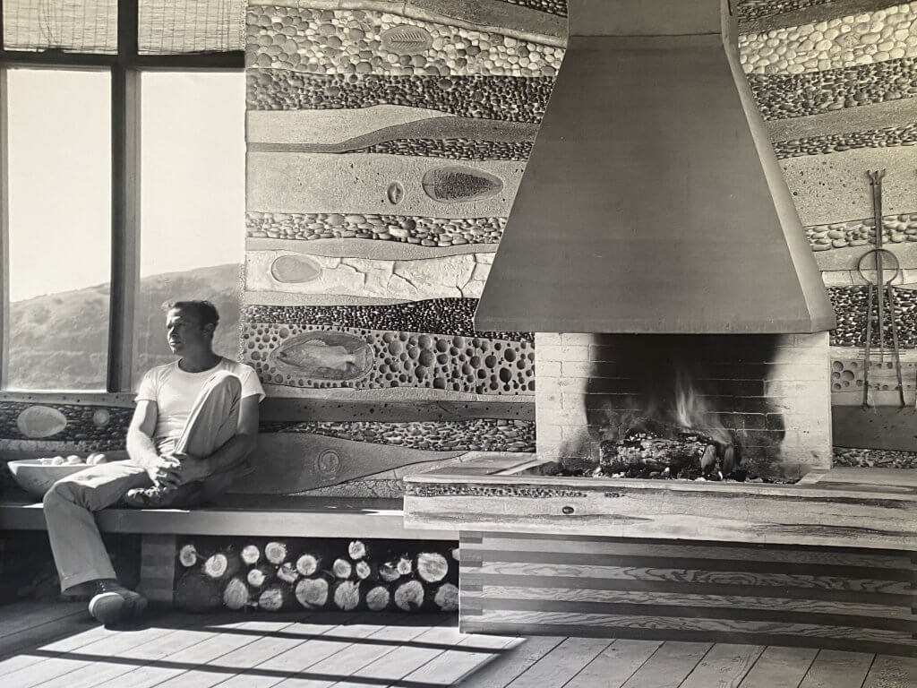 Black and white photo of artist Emile Norman who is sitting beside his fireplace in his home in Big Sur, circa 1956.