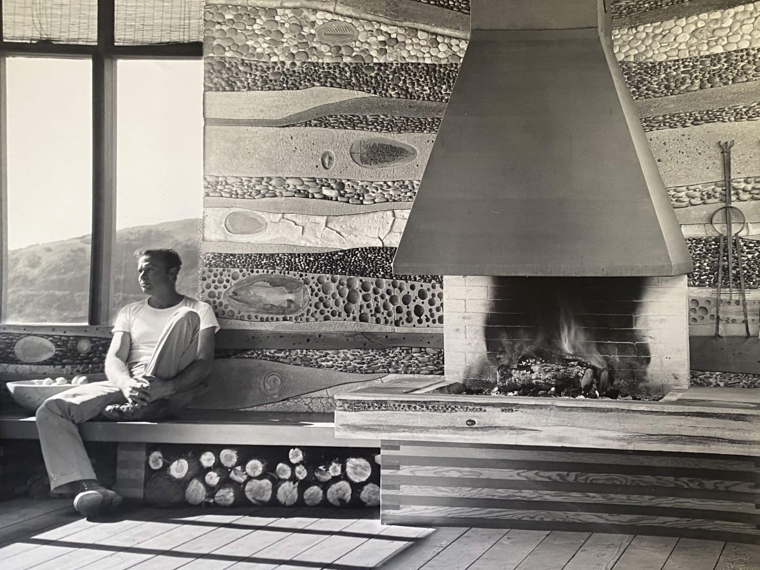 Black and white photo of artist Emile Norman who is sitting beside his fireplace in his home in Big Sur, circa 1956.